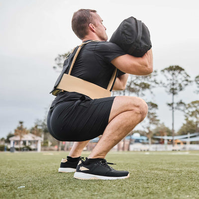 A person wearing GORUCK Men's Ballistic Trainers in Black, White, and Gold with a Reflective Gold Spearhead is doing squats with a weighted vest and sandbag on a grassy field.