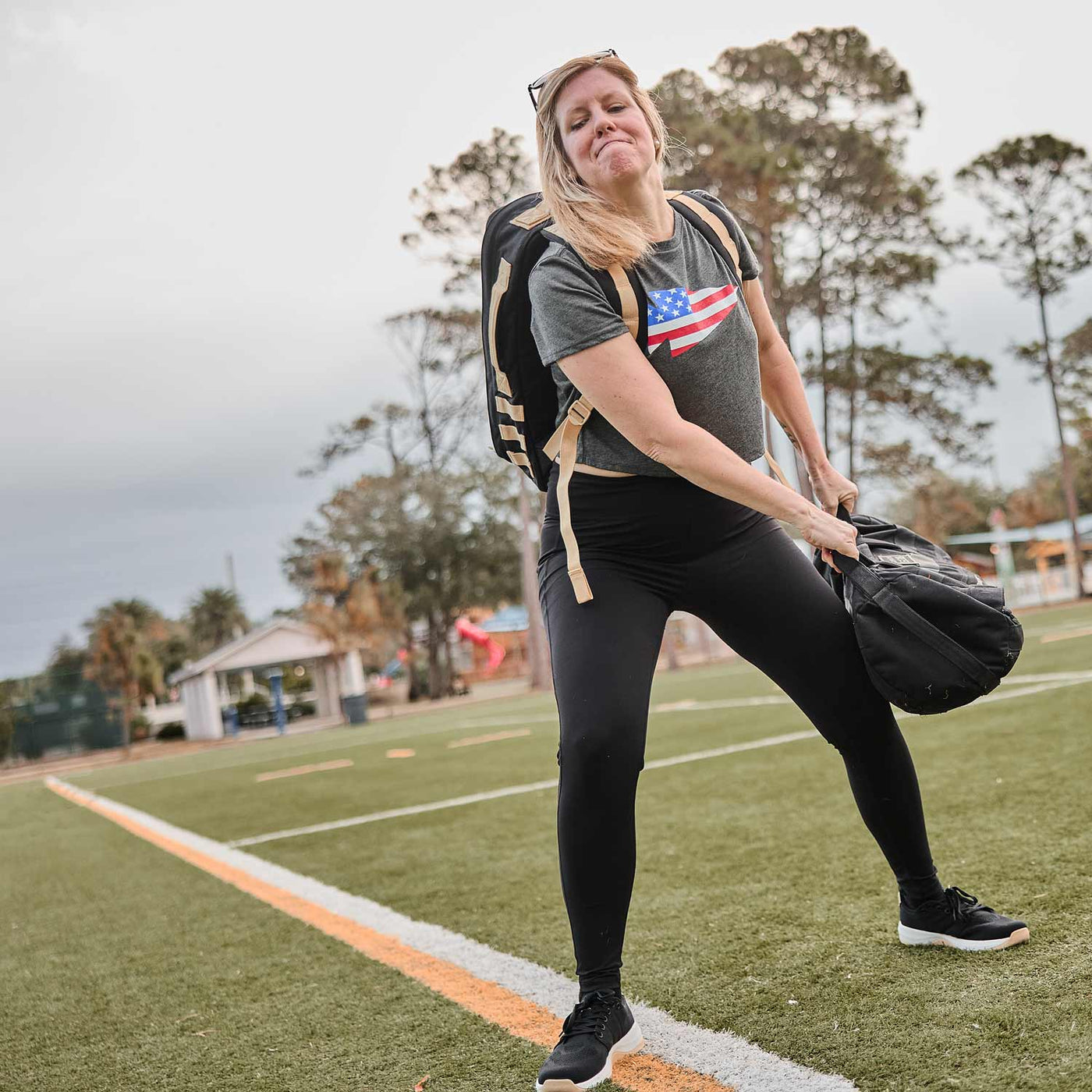 In a field surrounded by trees, someone showcases their strength by lifting a heavy bag while wearing a gray USA flag t-shirt and black leggings. They ensure stability with GORUCK's Women's Ballistic Trainers in Black, White, and Gold featuring the Gold Reflective Spearhead design.