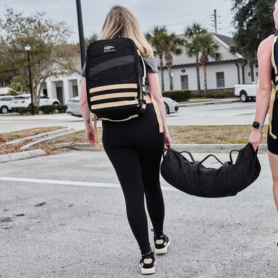 Two women carrying workout gear in a parking lot, one with a GORUCK backpack made of CORDURA® Ballistic Nylon and the other holding a sandbag, both confidently striding in their Women's Ballistic Trainers - Black + White + Gold W/ Gold Reflective Spearhead from GORUCK.