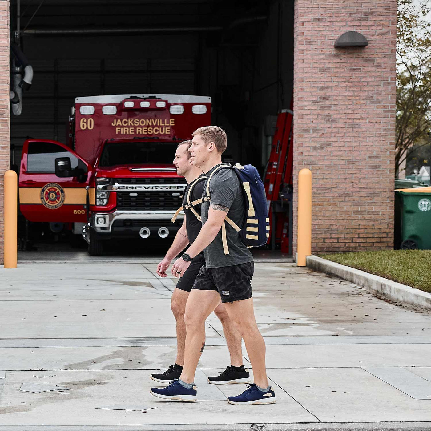 Two people wearing Men’s Ballistic Trainers from GORUCK, featuring rugged CORDURA® Ballistic Nylon, walk outside a fire station with fire trucks visible in the background.