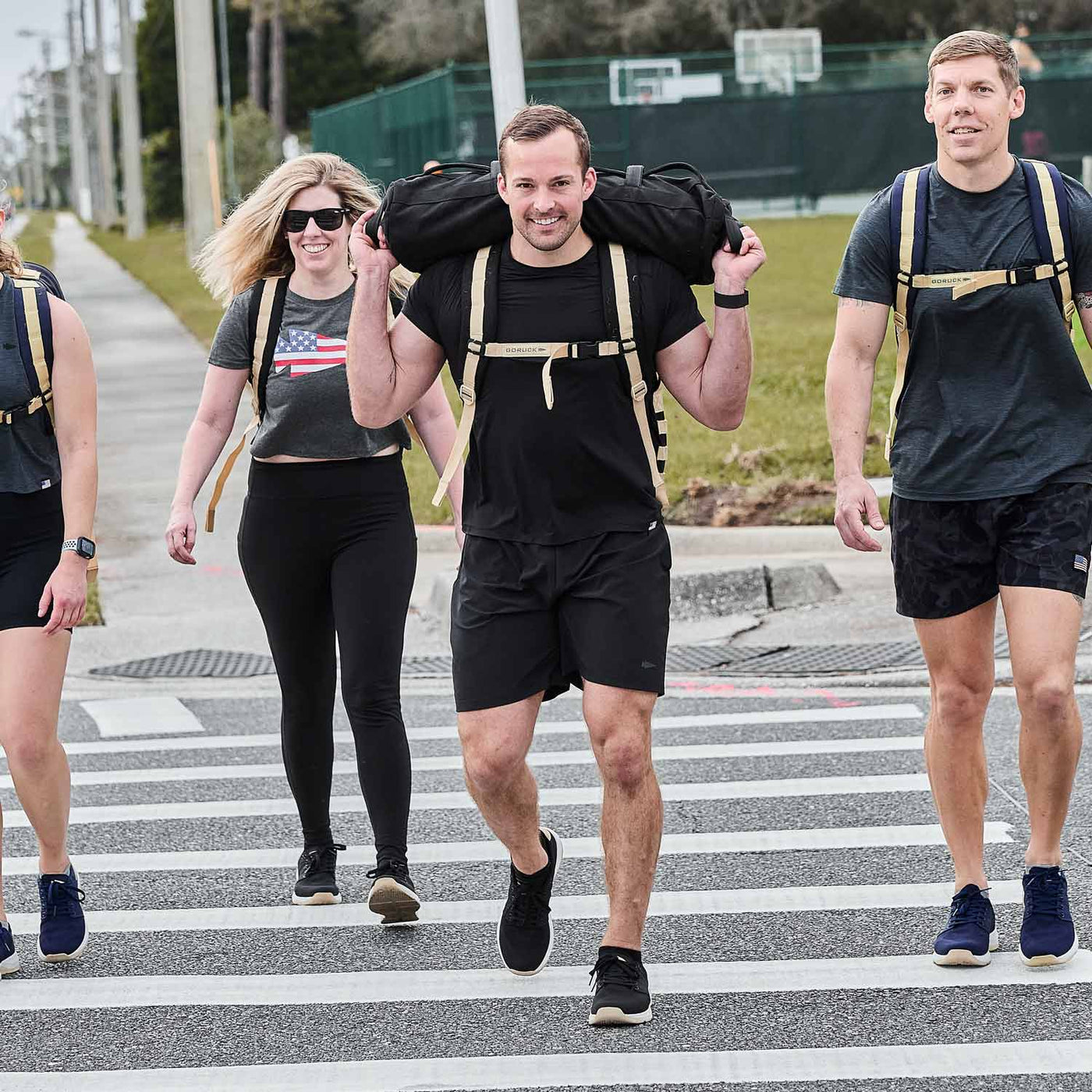 A group of people crosses the street, smiling and walking together with their backpacks, all wearing GORUCK’s Men’s Ballistic Trainers in Black, White, and Gold with a Gold Reflective Spearhead.