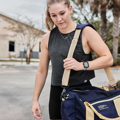 A woman with a smartwatch carries a GORUCK Kit Bag with shoulder strap in blue and tan, exuding the readiness of a Special Forces operative as she strides confidently outdoors.