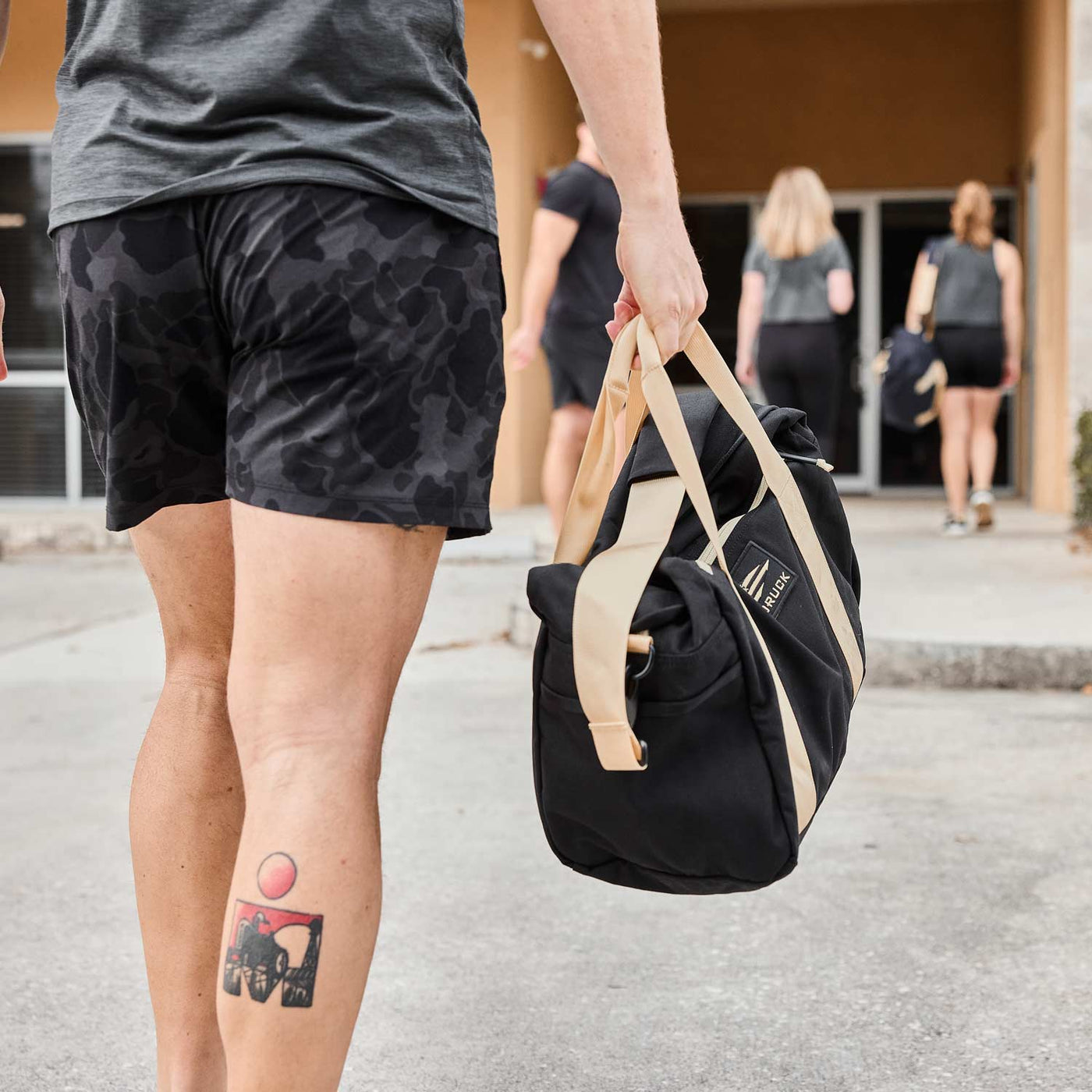 A tattooed individual carrying a GORUCK Kit Bag (Includes Shoulder Strap) walks toward a building alongside others dressed in athletic wear.