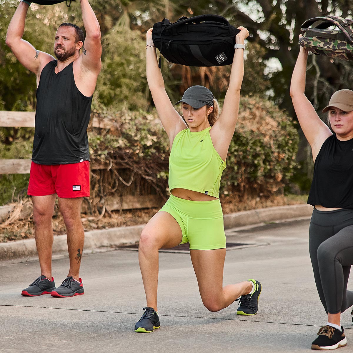 Three people exercise outdoors, holding weighted bags overhead. The center person sports a neon green outfit. On the left, someone in a black tank and red GORUCK Women’s Squat Shorts - ToughFlex exudes focus. To the right, another in high-waisted black leggings with a cap displays energetic determination, thanks to the comfort of ToughFlex Fabric.