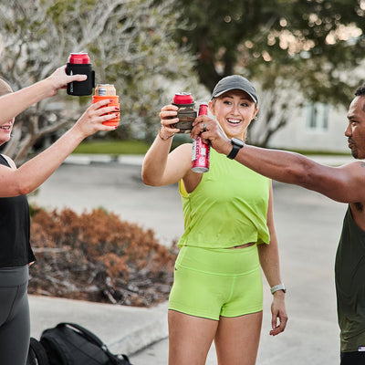 Three people are outside toasting with canned drinks, showcasing their athletic wear from GORUCK. One is wearing high-waisted Women’s Squat Shorts in bright green, and another sports a stylish cap. Thanks to the comfort of ToughFlex fabric, they celebrate effortlessly, with trees and a building creating the perfect backdrop for their joyous moment.