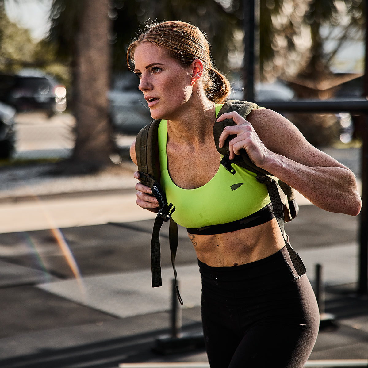 A woman in a vibrant green Power Bra - Toughflex from GORUCK pairs it with black leggings to jog outdoors in a park, with a weighted backpack on her back. Her GORUCK gear ensures she stays focused during her high-impact movements, while the trees and parked cars bask in the sunlight nearby.