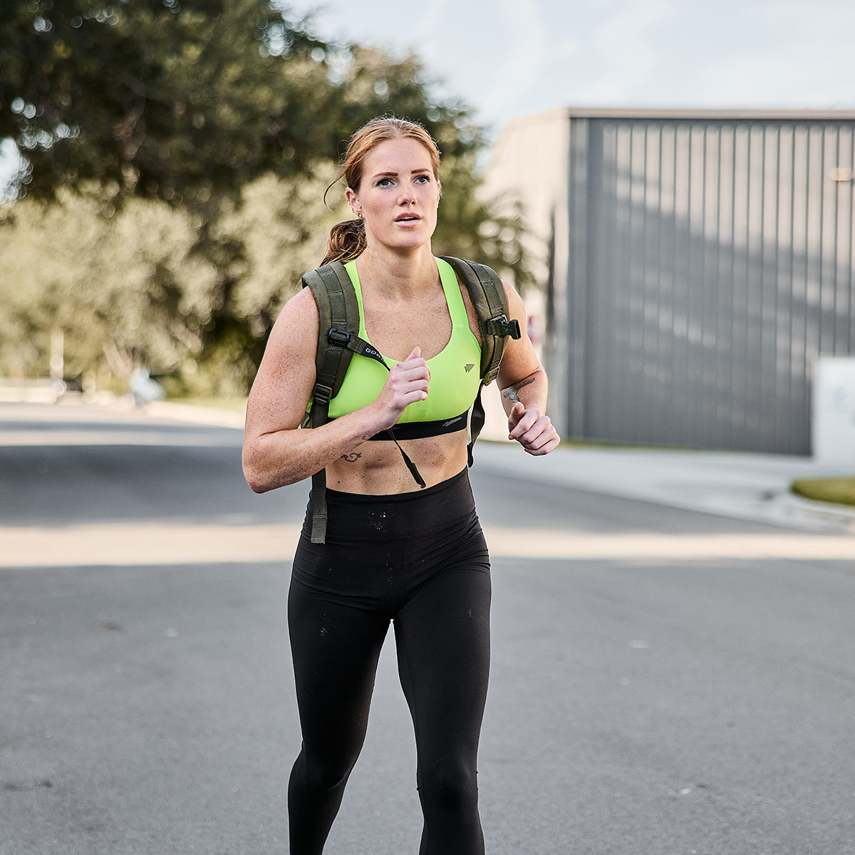 A woman jogging outdoors on a road exudes focus, effortlessly tackling high-impact movements. She sports a green Power Bra - Toughflex from GORUCK, paired with black leggings and a backpack. Her long hair is tied back, and trees and a building provide the perfect backdrop to her run.