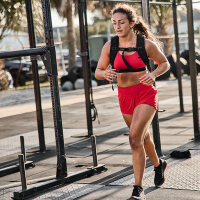 A woman jogs outdoors in a gym setting, dressed in squat-proof red sports apparel, including the GORUCK Women’s Training Shorts made from ToughStretch fabric, paired with a matching sports bra. She completes her outfit with a weighted vest and black running shoes, surrounded by fitness equipment under the shining sun.