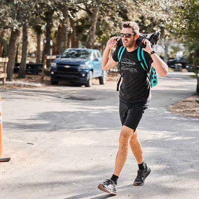 A man rucking outdoors with the Rucker 4.0 backpack and yoga mat, wearing sunglasses and casual sportswear.