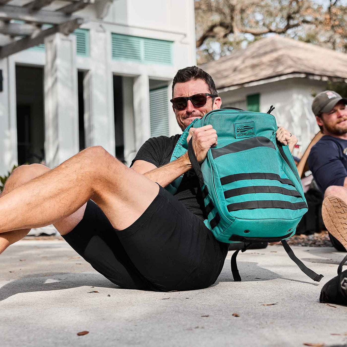 A man in sunglasses and a black shirt sits on the ground with a Rucker 4.0 teal backpack next to him, ready for a day of rucking adventure.