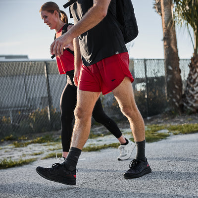 Two people stroll outdoors. One wears a shirt made from ToughStretch fabric for ultimate comfort along with red shorts known as Men’s Ranger Panties by GORUCK, while the other sports a red top and black leggings. They walk on a pavement lined with palm trees beside a chain-link fence under the bright daylight.