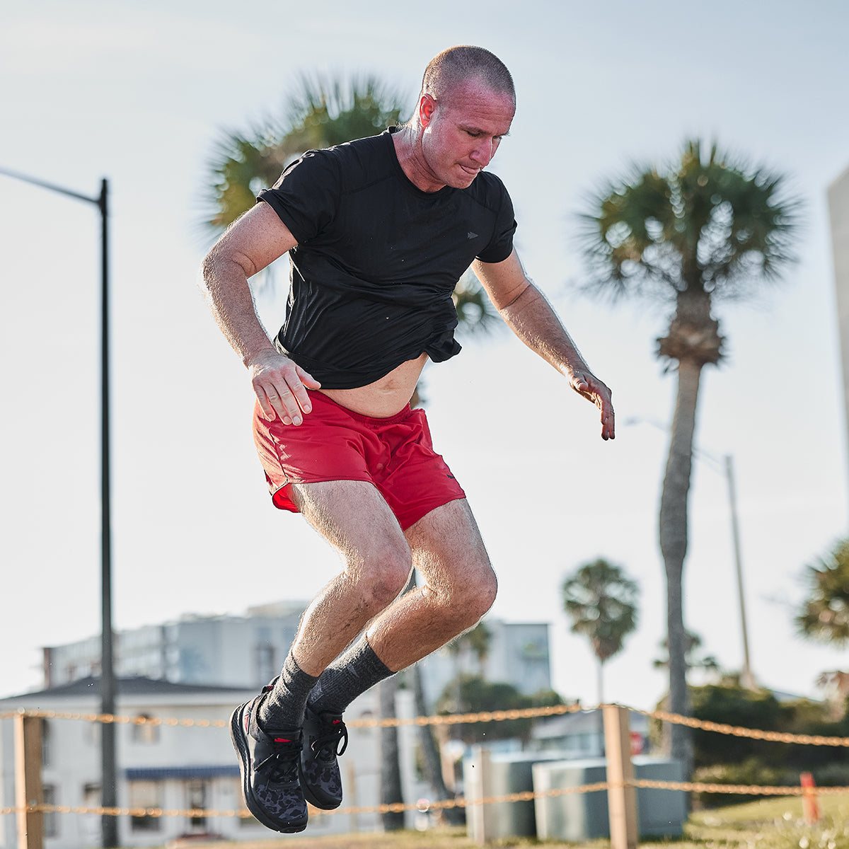 A person in a black shirt and GORUCK's Men's Ranger Panties - ToughStretch is mid-jump during an outdoor workout, set against a backdrop of palm trees and a clear sky.