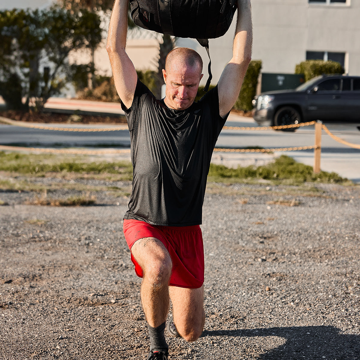 A man wearing a black T-shirt and GORUCK Men’s Ranger Panties with ToughStretch fabric performs an outdoor workout, lifting a heavy bag overhead while kneeling on one knee on the gravel. A building and parked car are visible in the background.