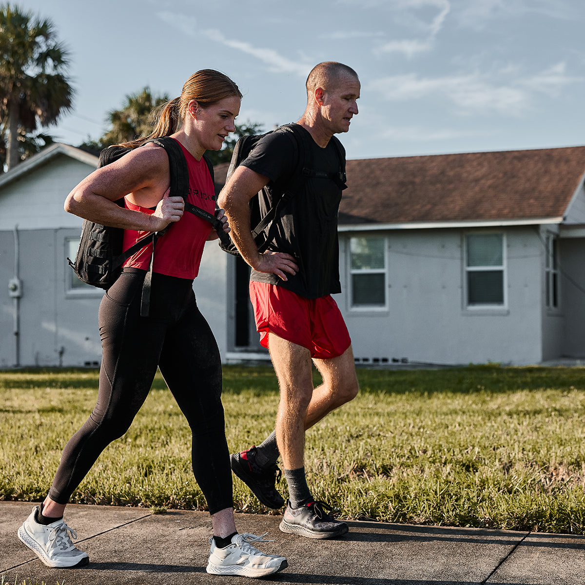 A man and woman, donned in GORUCK Men’s Ranger Panties crafted from ToughStretch fabric, walk briskly along a sidewalk in a residential area. The sky is clear, and a house with a red roof is visible behind them, exemplifying the gear's durability and backed by Scars Lifetime Guarantee.