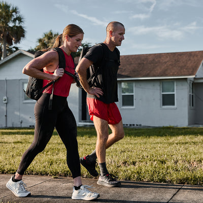 A man and woman, donned in GORUCK Men’s Ranger Panties crafted from ToughStretch fabric, walk briskly along a sidewalk in a residential area. The sky is clear, and a house with a red roof is visible behind them, exemplifying the gear's durability and backed by Scars Lifetime Guarantee.