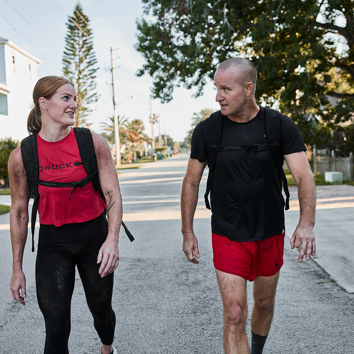 Two people stroll down a sunny street, sporting athletic gear and backpacks. The woman in a red top and black leggings smiles at the man in GORUCK Men’s Ranger Panties - ToughStretch shorts. Trees and houses line the background, adding a charming touch to their invigorating walk.