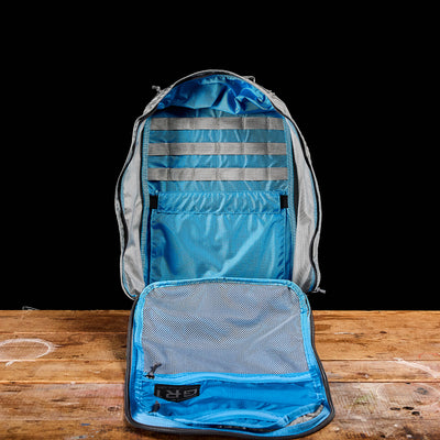 The GR1 USA - Ripstop ROBIC® backpack by GORUCK, featuring an ultra lightweight and water-resistant blue and gray design, sits open on a wooden table against a black background.