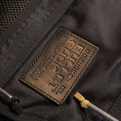 Close-up of a black fabric bag featuring a brown leather patch inscribed with "Bullet Heritage - Built by SCARS." The patch is sewn onto this retro canvas rucksack from GORUCK, with part of a zipper visible and adorned with a paracord pull.