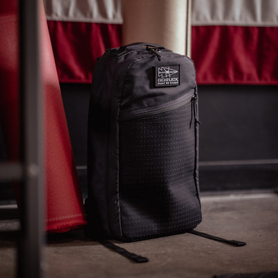 A black Bullet Heritage - Built by SCARS backpack from GORUCK is positioned upright on a concrete floor, with a red punching bag and large American flag blurred in the background. Reminiscent of a retro canvas rucksack, it showcases a subtle grid pattern along with its patch logo on the top front.