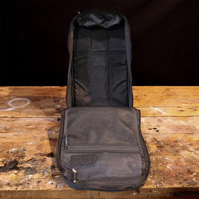 The Bullet Heritage - Built by SCARS, crafted by GORUCK, lay open on a wooden surface. Its black design echoes a retro canvas rucksack style, showcasing a mesh interior pocket and zippered compartments. The rustic wood beneath it is adorned with paint stains and signs of age.