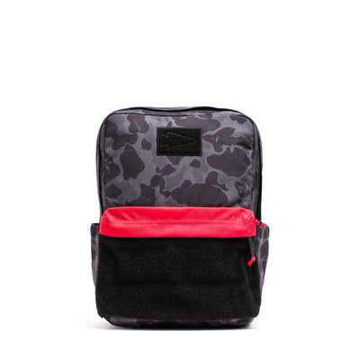 The KR1 2.0 - Kid Ruck by GORUCK is a durable kids backpack, showcasing a black and gray camo design with reinforced stitching, complemented by a vibrant red front pocket and a sturdy black bottom section.