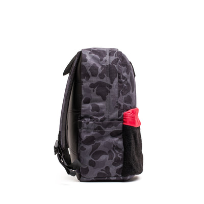 Side view of the KR1 2.0 - Kid Ruck by GORUCK, a gray camouflage kids backpack with reinforced stitching and a red towel tucked into the side pocket.