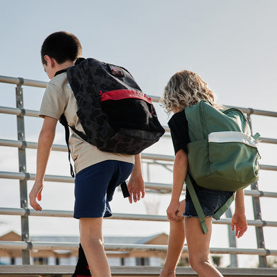 Two children with their GORUCK KR1 2.0 - Kid Rucks climb the metal bleachers under a sunny sky, their backpacks featuring reinforced stitching to handle any adventure.