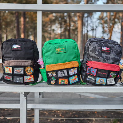 Three vibrant GORUCK KR1 2.0 - Kid Ruck backpacks adorned with assorted patches are displayed on a bench in an outdoor setting, highlighting their robust design and reinforced stitching.