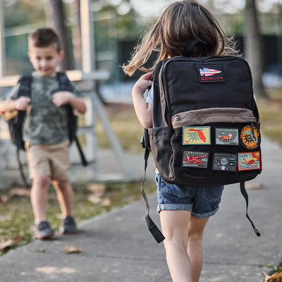 Two young children with backpacks walk on a sidewalk; in the foreground, the girl is carrying a GORUCK KR1 2.0 - Kid Ruck, which features reinforced stitching and a patch-decorated design.