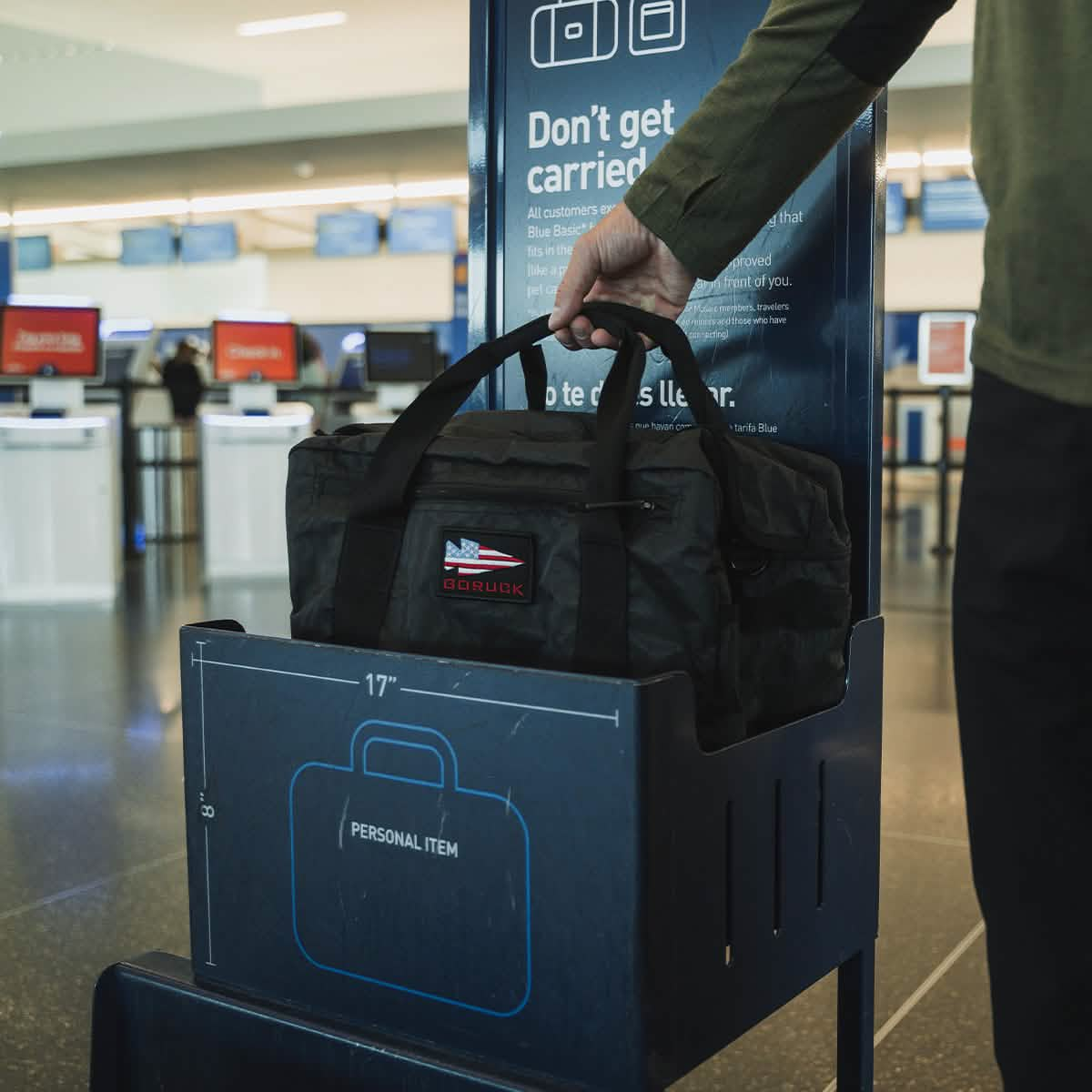 A traveler places a sleek black Kit Bag - Dyneema, made by GORUCK, into a personal item size checker at the airport. Next to it is a sign outlining luggage dimension guidelines, while in the background, check-in kiosks and counters are bustling with travelers.