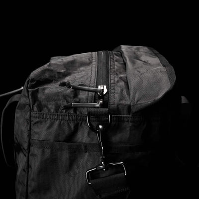 Close-up of the Kit Bag - Dyneema by GORUCK, reminiscent of an Aviator’s Kit Bag, set against a dark background. This black fabric bag boasts a prominent zippered closure and a sturdy strap accented with sleek metal hardware. The Dyneema material showcases its texture with a slightly crumpled appearance.