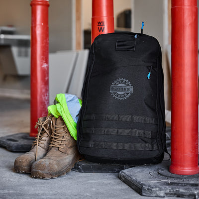 The GR1 USA - GORUCK X mikeroweWORKS backpack showcases American craftsmanship, leaning against red poles, surrounded by brown boots and green safety gear in an industrial setting that epitomizes hard work and dedication.