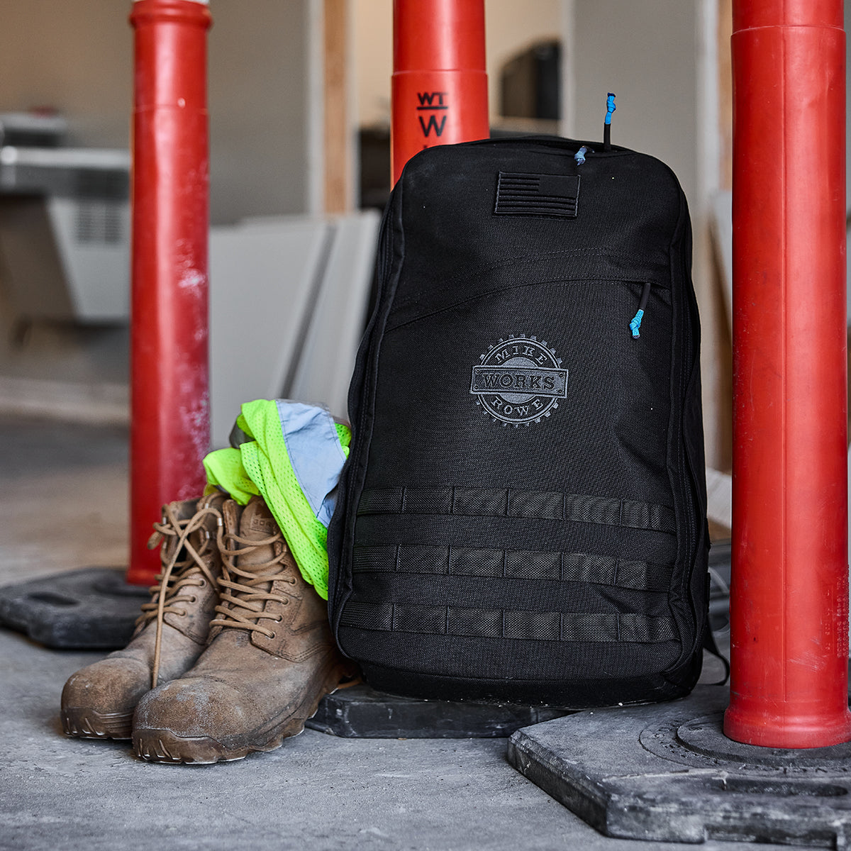 The GORUCK X mikeroweWORKS Bundle, featuring a black backpack and sturdy work boots, contrasts with vibrant neon clothing near red poles at a construction site, highlighting the rugged professions backed by the mikeroweWORKS Foundation.