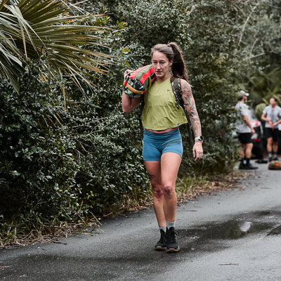 A woman in athletic wear, including Women's Squat Shorts - ToughFlex, carries a sandbag on her shoulder along a wet path amidst greenery.