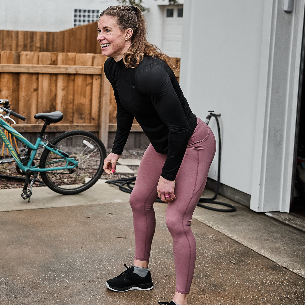 A woman in a black top and pink Women’s Training Leggings Pocket - ToughFlex exercises outdoors by a green bicycle, confidently showcasing her squat-proof style with every move.