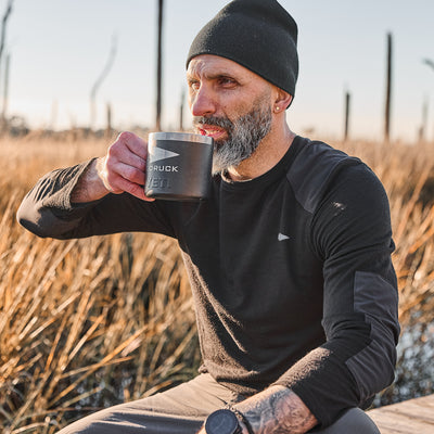 A man in a black beanie and shirt relishes his mug outdoors among tall grass, with his attire reflecting the rugged elegance of a Men’s Commando Long Sleeve - Merino Wool sweater, enhancing the serene atmosphere.