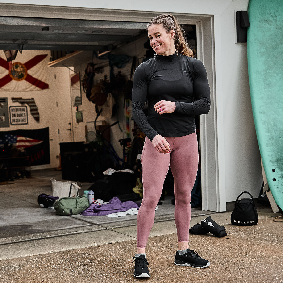 A woman in athletic wear, wearing the Women’s Long Sleeve Mock Neck - ToughMesh, stands smiling outside a garage surrounded by surfboards and workout gear. The outfit's moisture-wicking properties provide comfort as she embraces her active lifestyle.