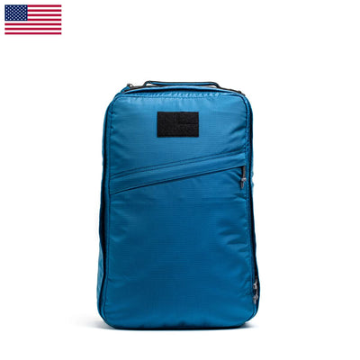 The ultra lightweight GR1 USA - Ripstop ROBIC® travel backpack by GORUCK, featuring a blue design with a diagonal zip pocket, is showcased against a white background with a small U.S. flag in the corner, highlighting its water-resistant capabilities.