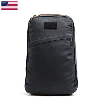 The GR1 USA - Heritage by GORUCK is a black tactical backpack that brings to mind a classic rucksack design. It prominently displays a rectangular patch in the center and confidently stands upright against a plain background. The backpack features a zippered front pocket and sturdy handles, complemented by an American flag in the top left corner for a touch of patriotic flair.