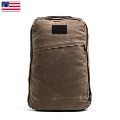 A brown GR1 USA - Heritage backpack from GORUCK, with a rectangular design, includes a zippered front pocket. Crafted from durable material, it features a small black patch near the top and proudly displays the US flag in the upper left corner against a plain white background, enhancing its classic appeal.