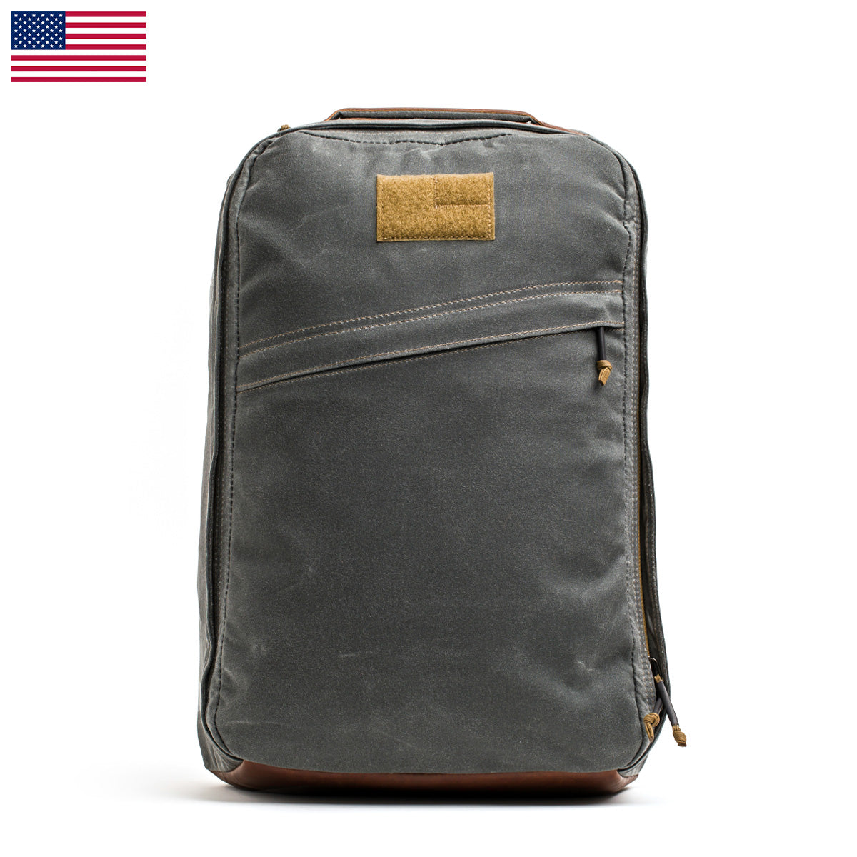 The GR1 USA - Heritage, a GORUCK product, is displayed against a white background. It highlights its gray design with a leather base and accents. The bag includes a front pocket with a zipper and bears a small brown patch on the top front. An American flag is visible in the top left corner.