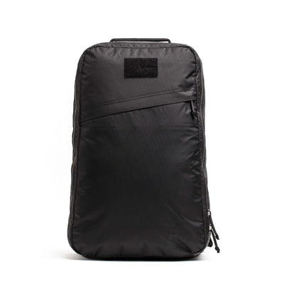 The GR1 USA - X-PAC by GORUCK is a black minimalist rucksack designed with a sleek look, featuring a subtle diagonal zipper pocket on the front and smooth, waterproof X-PAC texture. It includes a visible top handle and a side-running main compartment zip.