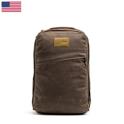 The GR1 USA - Heritage by GORUCK is a brown rucksack with a minimalist design, crafted from waxed canvas. It features a small front pocket and a tan patch, with the subtle USA flag in the top left corner, capturing the essence of the GR1 style. The bag is presented against a white background.