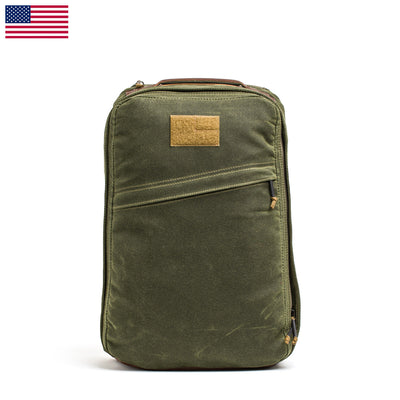 The GR1 USA - Heritage backpack by GORUCK is a green heritage bag with a tan accent on the front, made from waxed duck canvas and featuring multiple zippers. An American flag in the top left corner signifies its American origin. The backpack is photographed upright against a pristine white background.