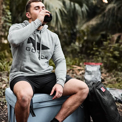 A person in a GORUCK Poly-Blend pullover sits on a cooler outdoors, sipping from a bottle. Next to them is a black backpack featuring the GORUCK spearhead design, reflecting its Special Forces heritage, while a wooded area stretches into the background.