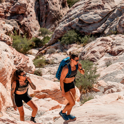 Two individuals are ascending rocky terrain with GORUCK Rucker 4.0 backpacks, surrounded by rugged cliffs and sparse greenery.