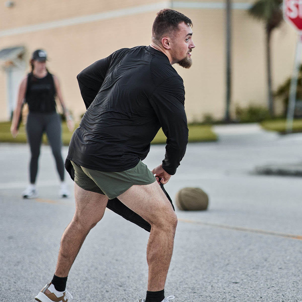 A man wearing a GORUCK Men's Long Sleeve Performance Tee in ToughMesh fabric, paired with green shorts, is exercising outdoors by pulling a heavy bag along the pavement. In the background, a woman is seen standing in leggings and a cap. A stop sign can be spotted in the distance, evoking imagery of trails in the Italian Alps.