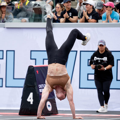 A competitor, clad in black leggings and a brown top, displays impressive skills in a handstand walk during the event. With the audience's applause filling the air, the camera operator documents every moment while the GORUCK Women's Ballistic Trainers - Lunar Rock + Gum with Silver Reflective Spearhead ensure stability and support for the athlete.