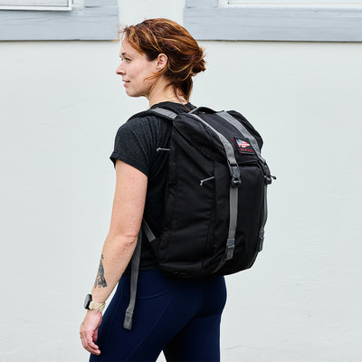 A person with a tattoo wearing the sleek and modern M23 - Cordura rucksack by GORUCK stands against a white wall.