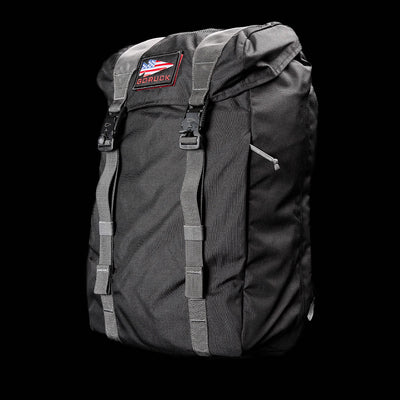 The GORUCK M23 - Cordura rucksack is designed with sturdy materials and includes front straps along with an American flag patch on the top flap, all presented against a sleek black background.